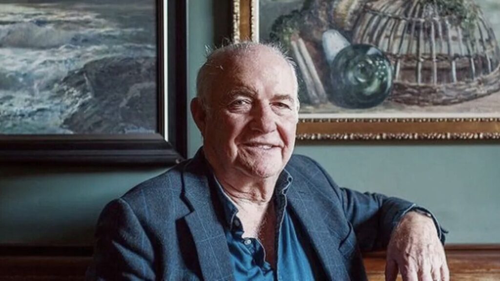 Rick Stein, an older man with short gray hair, wearing a blue blazer and an open-collared shirt, sits indoors in front of framed paintings, smiling slightly and resting one arm on a wooden surface.
