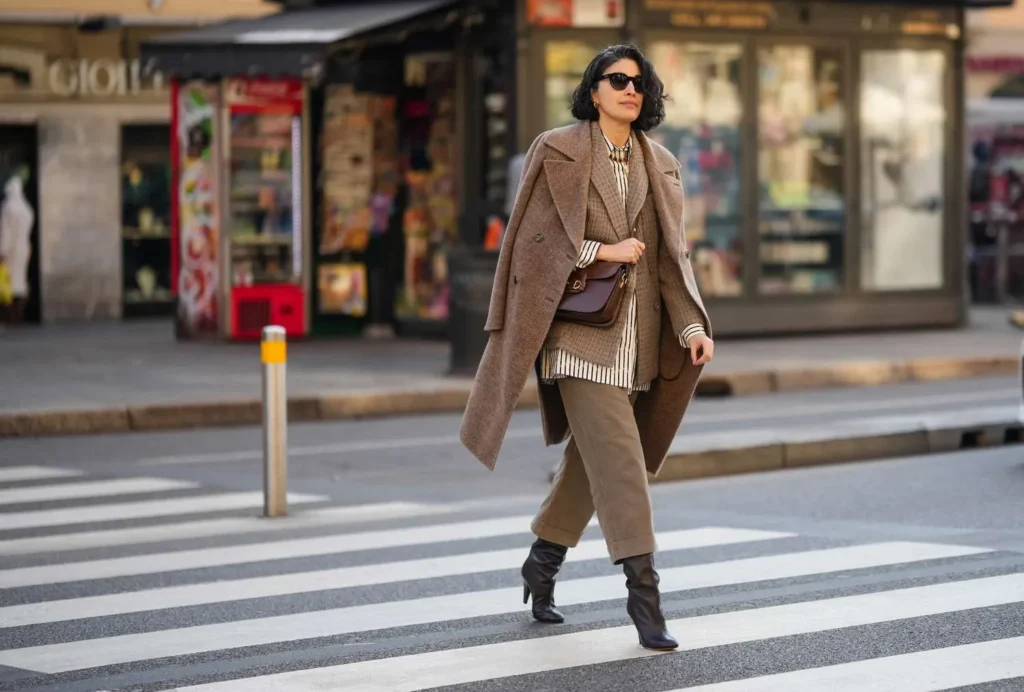 Woman wearing winter outfits with beige oversized coat, striped shirt, beige trousers and black ankle boots on city street