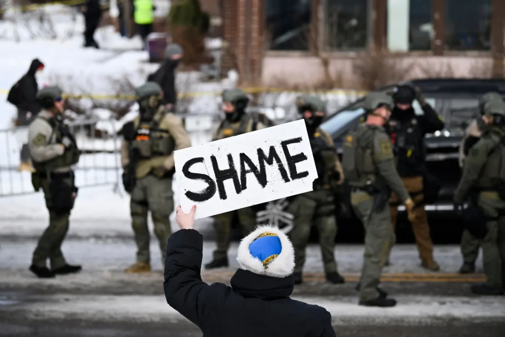 Protester holding 'SHAME' sign confronting ICE officer and federal agents in Minneapolis winter street demonstration